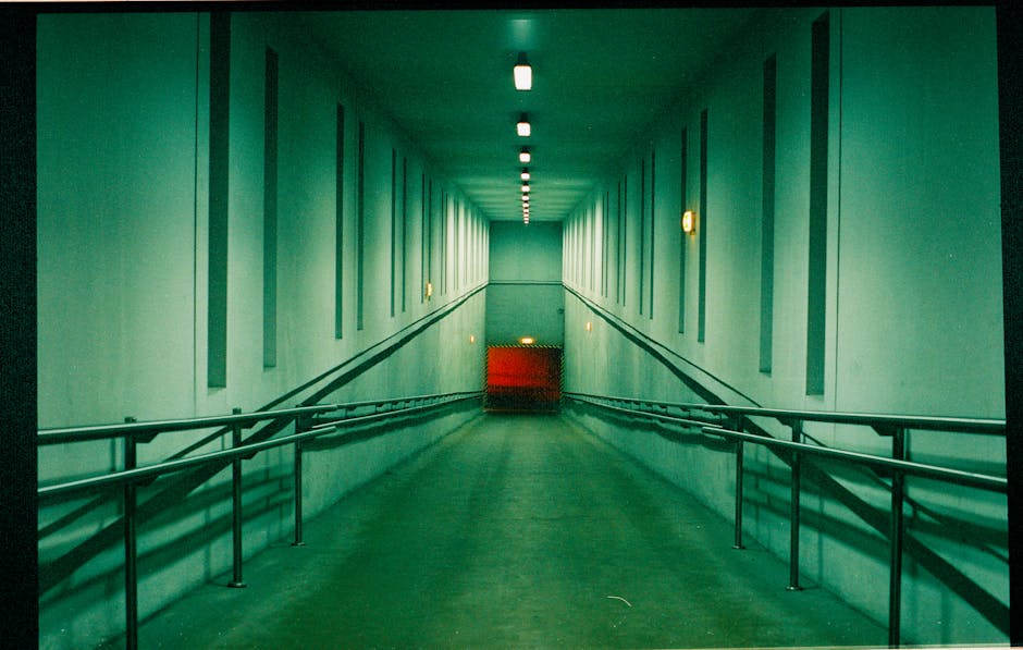 A long, empty corridor with dim lighting and an accessibility ramp leading to a red door.